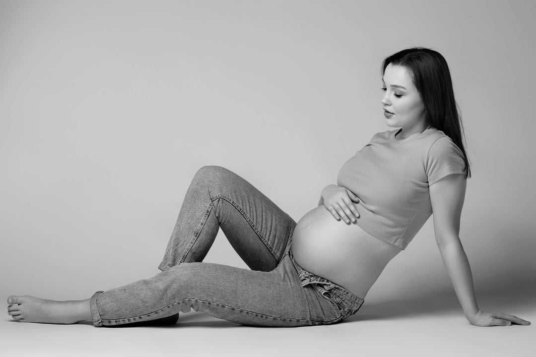 pregnant woman wearing postpartum shapewear and jeans, sitting on the floor and highlighting comfortable bodysuit support