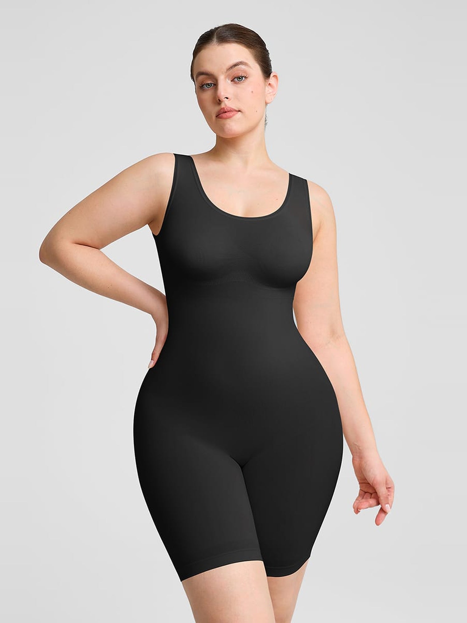 A woman modeling a black sleeveless shapewear bodysuit with built-in shorts, shown against a solid light gray studio background.