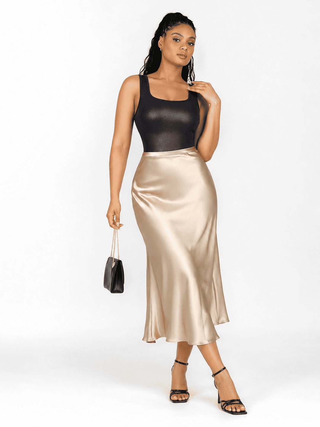 A model stands against a white studio background wearing a black sleeveless top and a shiny champagne-gold satin midi skirt, holding a small black handbag.