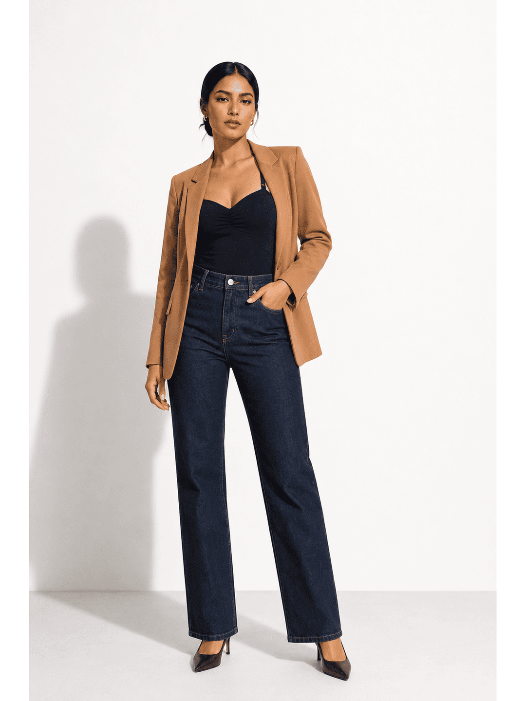 A woman stands against a white studio wall wearing a camel blazer, black camisole top, dark blue straight-leg jeans, and black pointed heels.