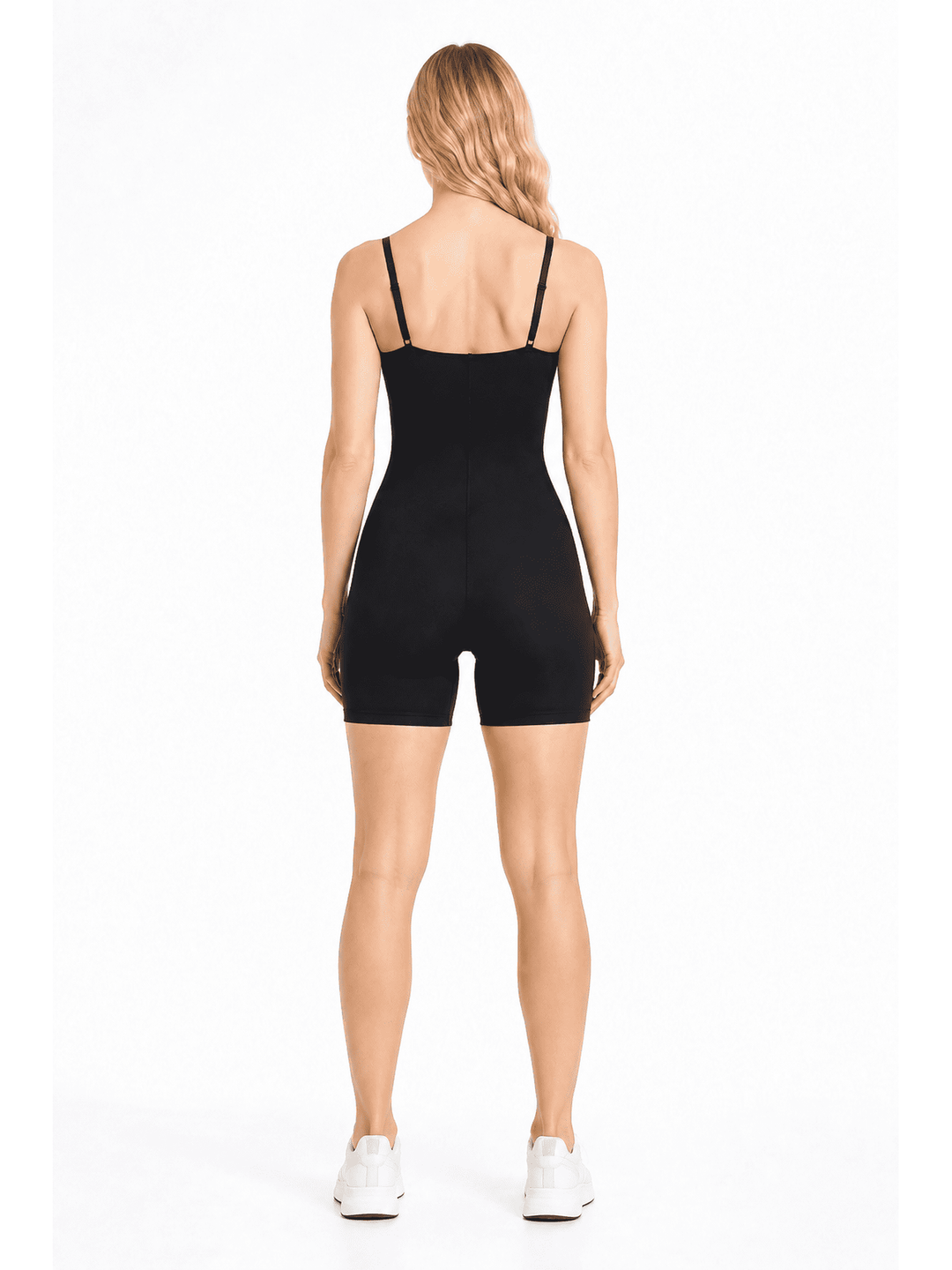 A woman viewed from the back wearing a black shapewear bodysuit with thin shoulder straps and fitted shorts, paired with white sneakers, against a plain white studio background.