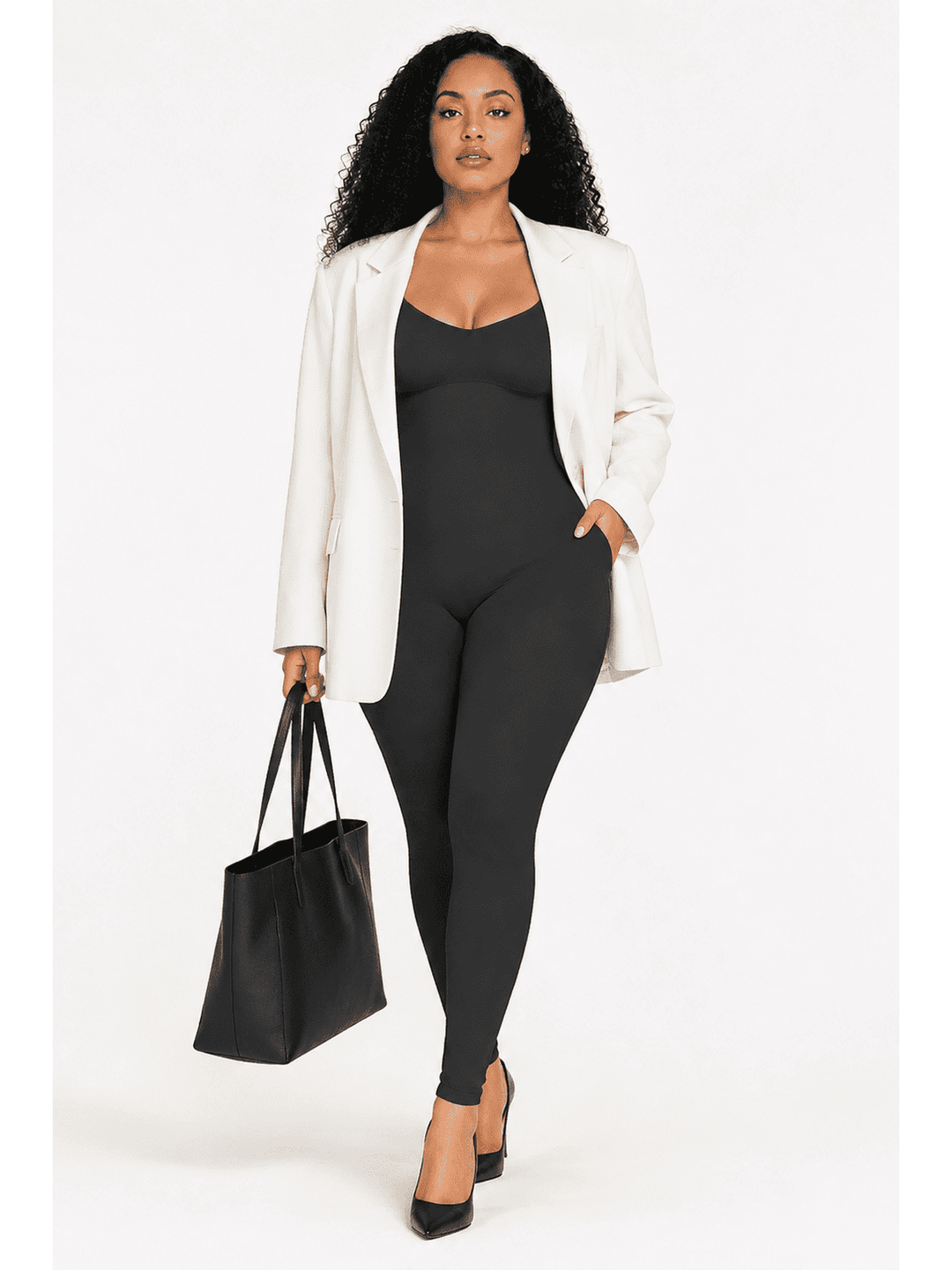 A woman modeling a black bodysuit with a white blazer, black heels, and holding a black tote bag against a plain white studio background.