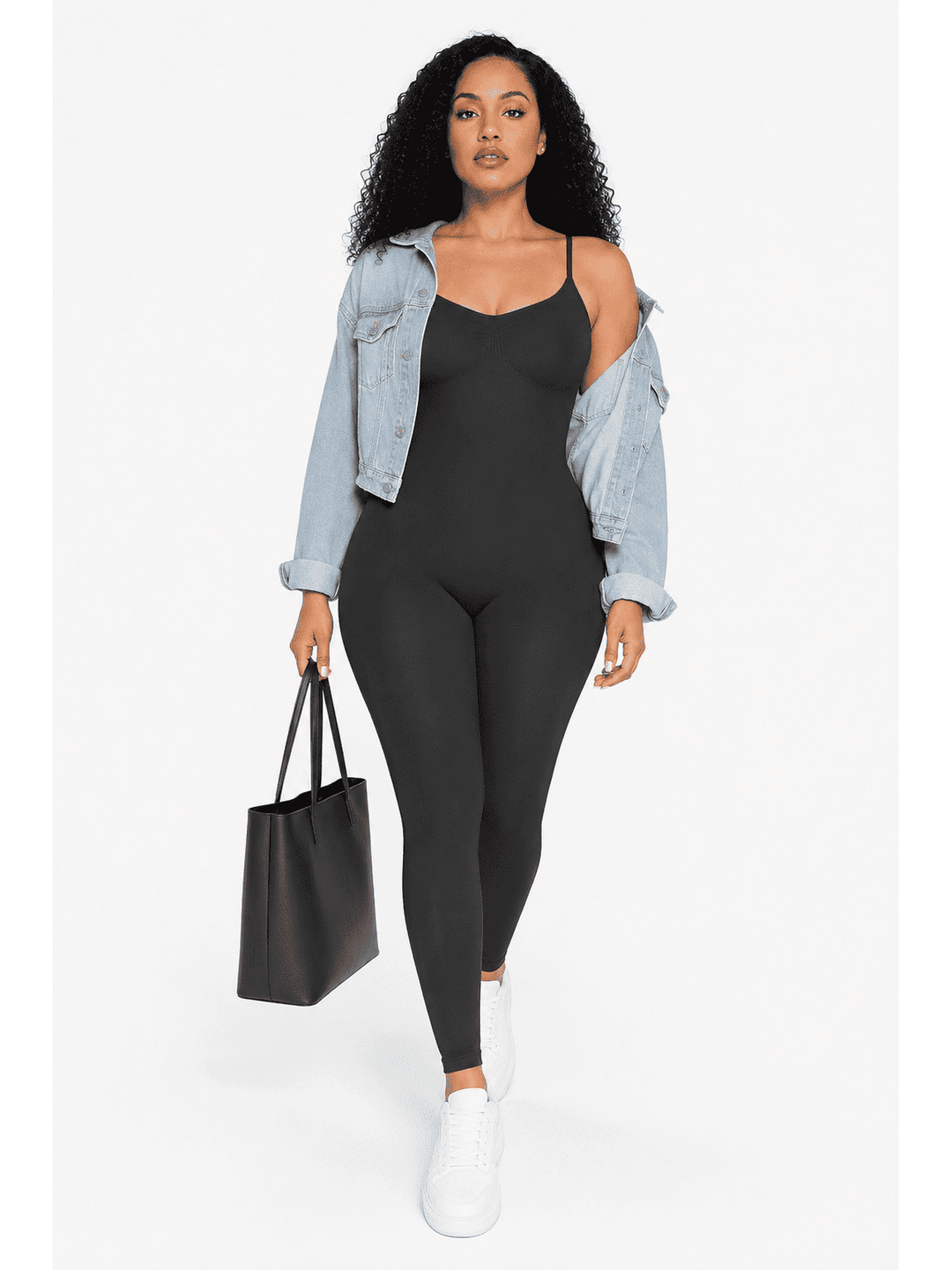 A woman modeling a black camisole jumpsuit with thin straps and full-length legs, styled with an open light-wash denim jacket, white sneakers, and a black tote bag against a plain white studio background.