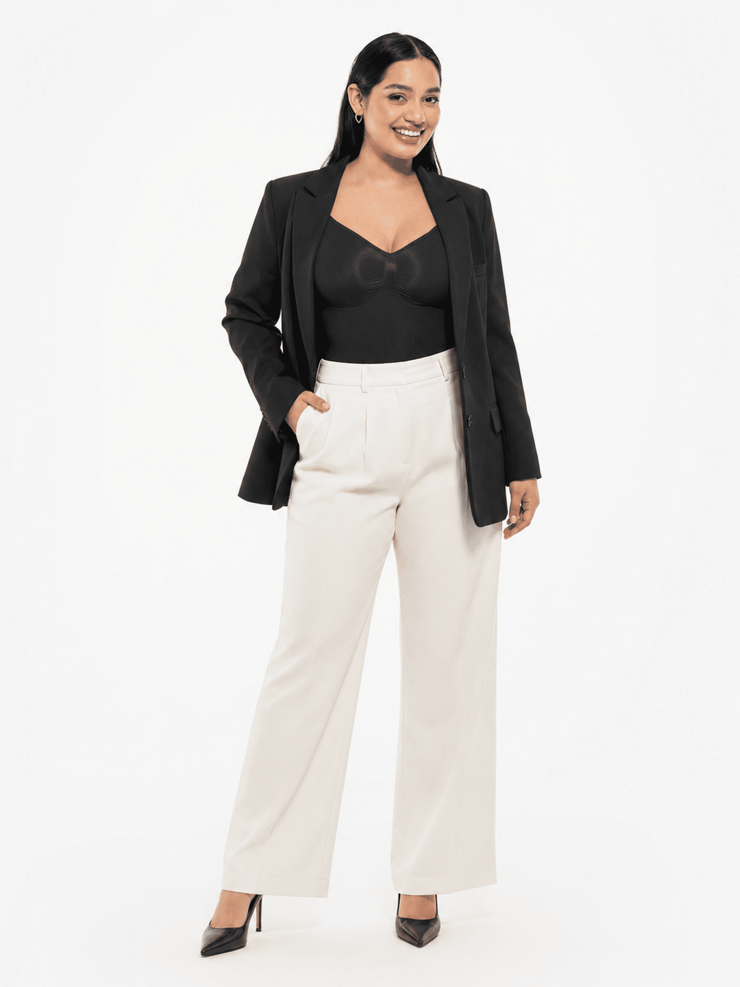 A woman modeling a black blazer over a black top with off-white wide-leg trousers and black heels, standing against a plain white studio background.