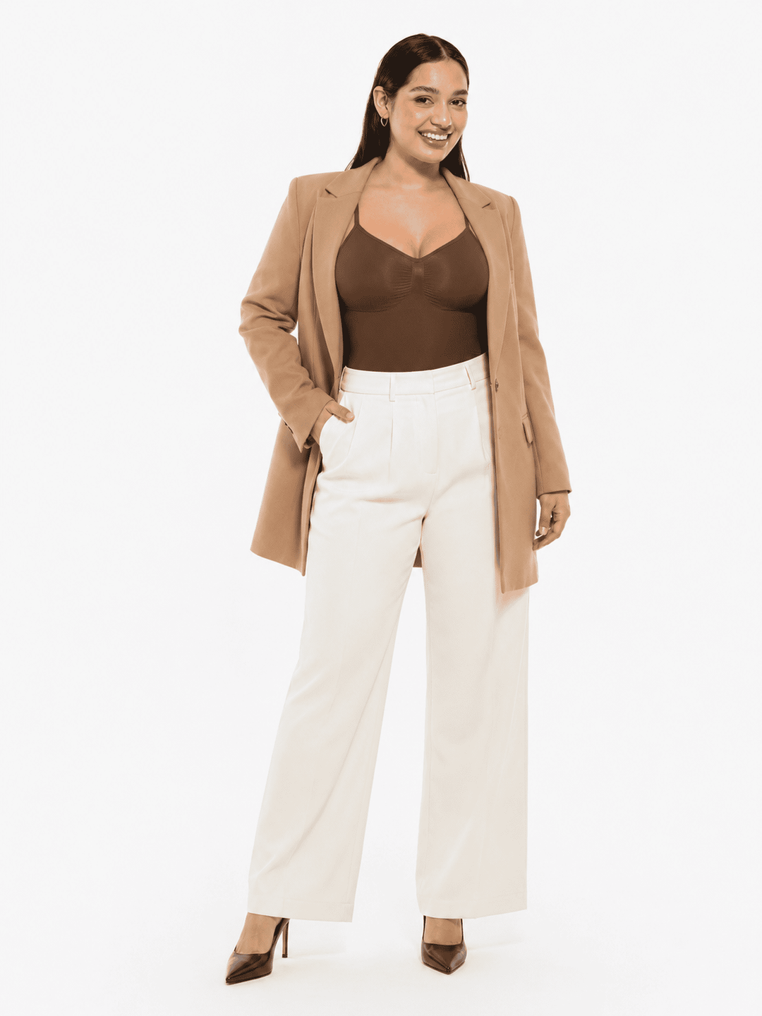 A woman modeling a tan blazer over a brown camisole and white wide-leg trousers, standing against a plain white studio background.
