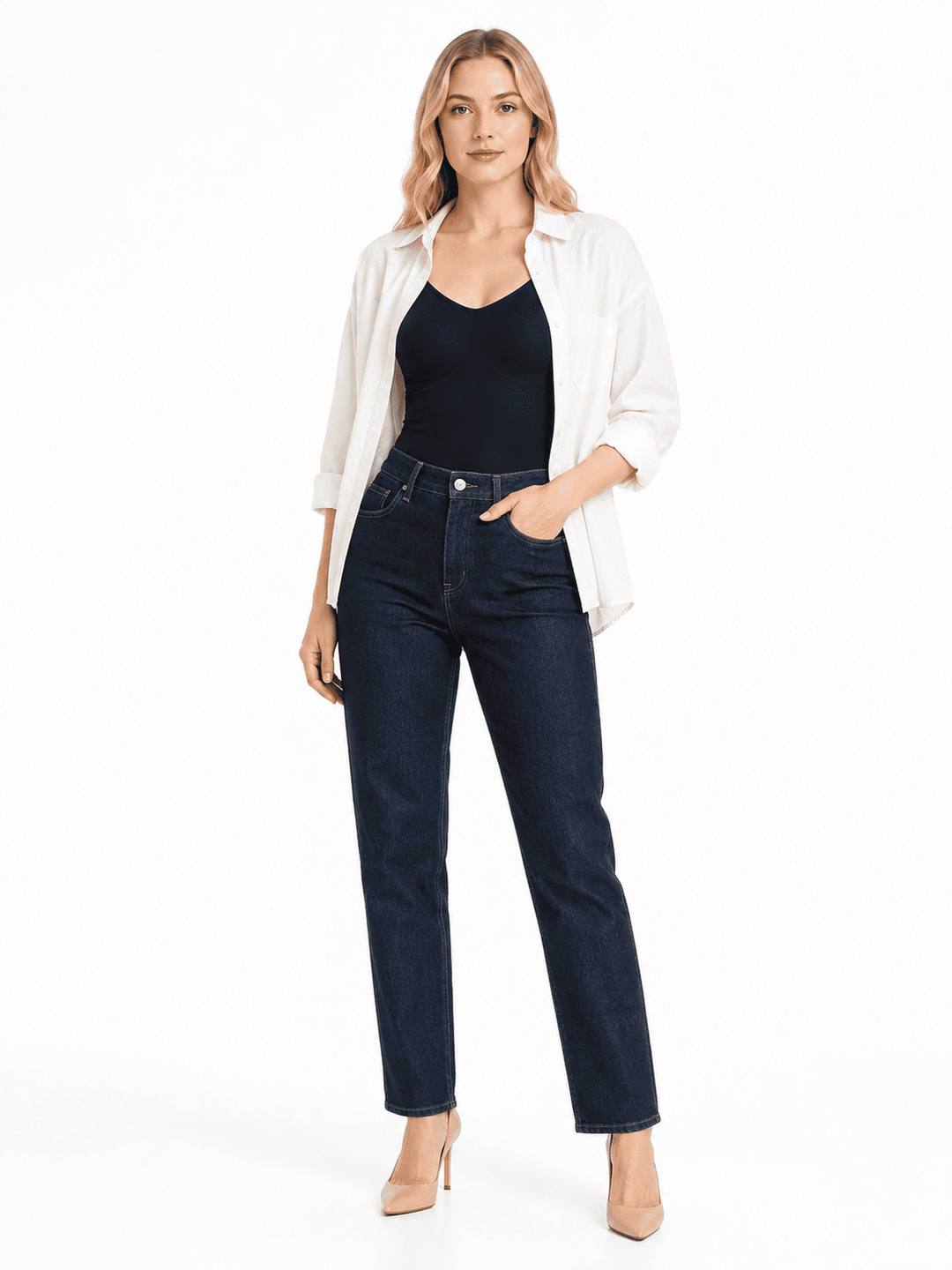 A woman modeling a pair of dark wash blue jeans with a black camisole and an open white shirt, standing against a plain white studio background.