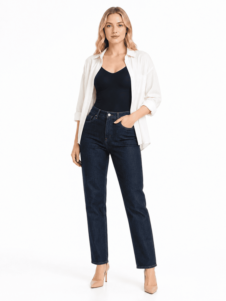 A woman modeling a pair of dark wash blue jeans with a black camisole and an open white shirt, standing against a plain white studio background.