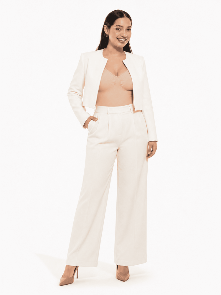 A woman modeling a matching off-white cropped blazer and wide-leg trousers set, styled with a nude bralette and beige heels against a clean white studio background.