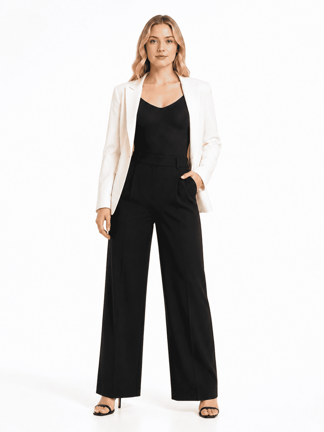 A woman modeling a black wide-leg jumpsuit with a black camisole underneath, styled with an open white blazer and black heeled sandals against a plain white studio background.