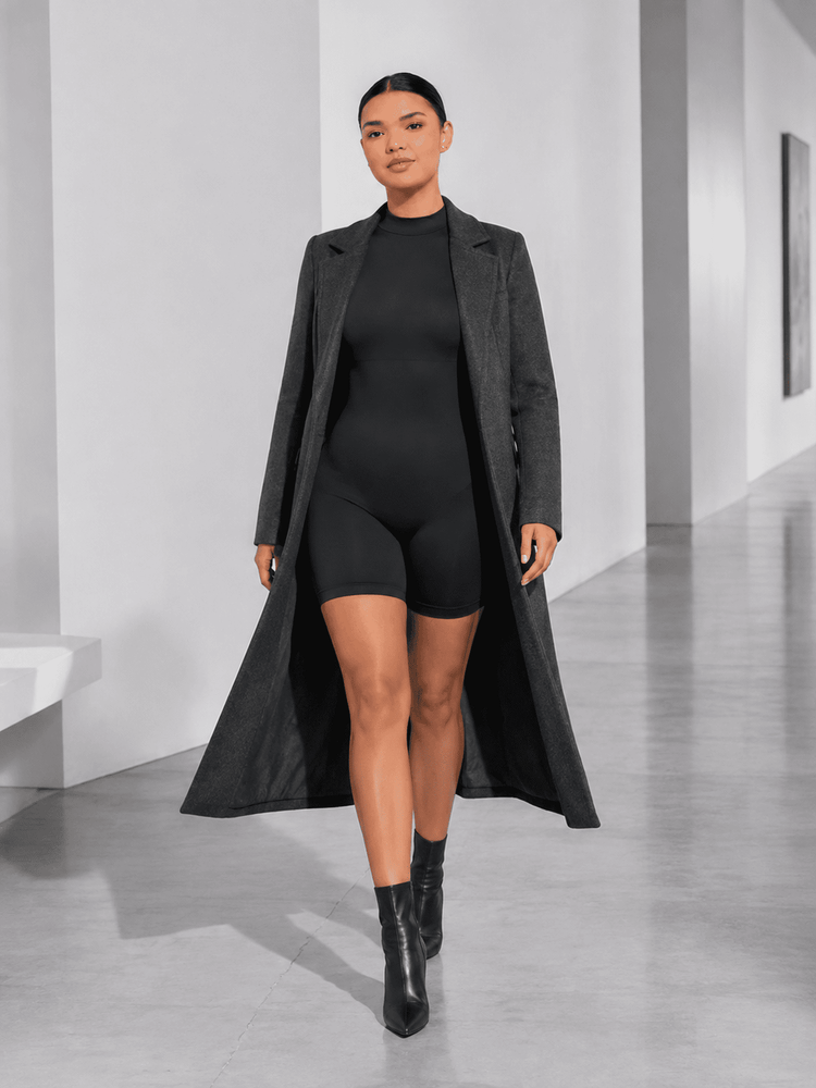 A woman walks down a minimalist white corridor wearing a long charcoal gray open-front coat over a black bodysuit and black shorts, paired with black ankle boots.