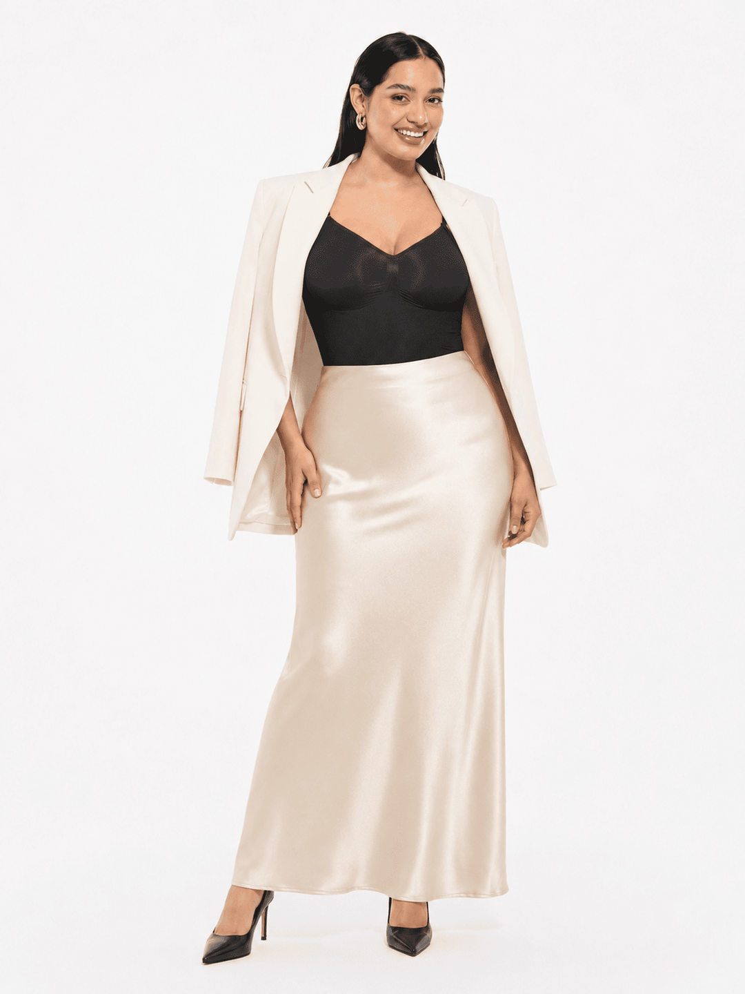A woman modeling a two-piece outfit featuring a black sweetheart-neckline camisole top and a high-waisted, floor-length satin slip skirt in champagne beige, styled with an open white blazer and black heels against a white studio background.