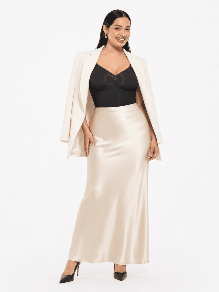 A woman modeling a two-piece outfit featuring a black sweetheart-neckline camisole top and a high-waisted, floor-length satin slip skirt in champagne beige, styled with an open white blazer and black heels against a white studio background.