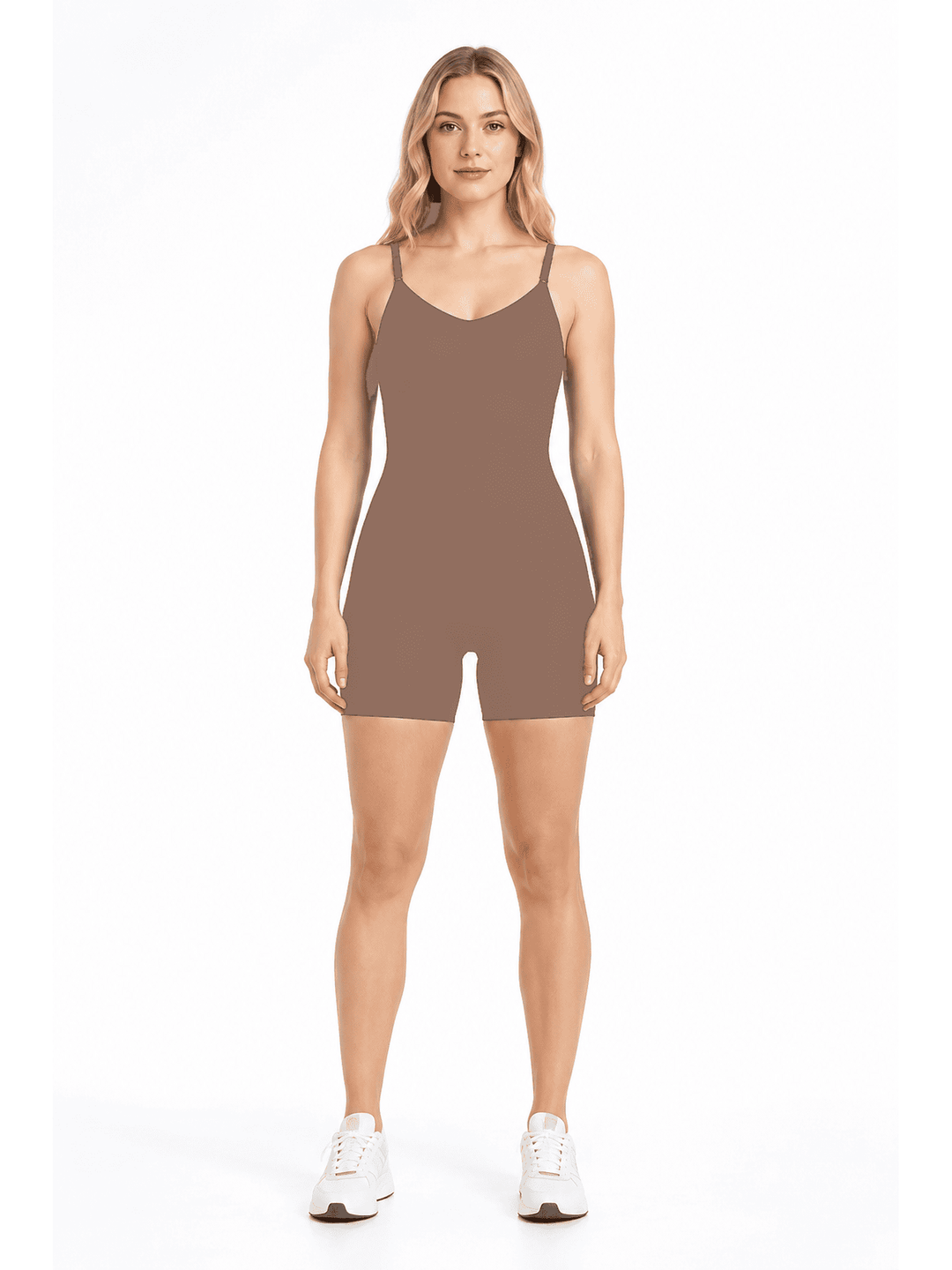 A woman modeling a sleeveless, short-biased athletic romper in a solid taupe-brown color, paired with white sneakers, against a plain white studio background.