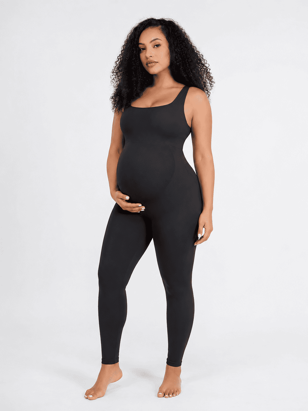 A pregnant model with curly black hair wearing a black maternity bodysuit and leggings, standing against a plain white studio background.