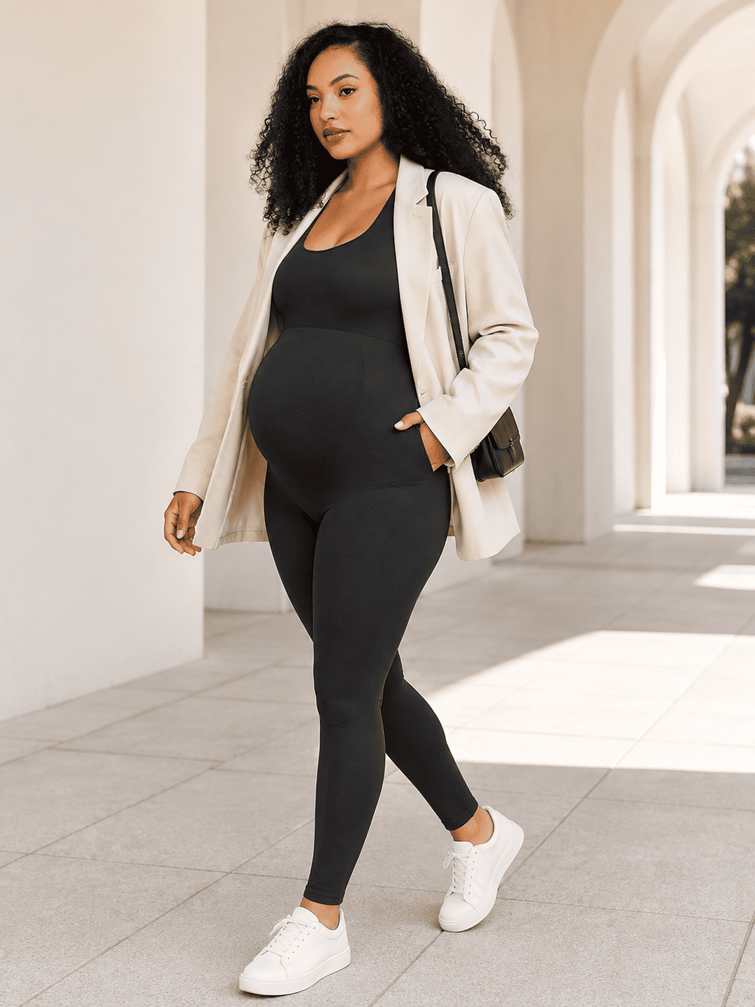 A pregnant woman wearing a black maternity bodysuit and leggings, styled with an open cream blazer, white sneakers, and a black shoulder bag, walking in a sunlit outdoor corridor with arches.