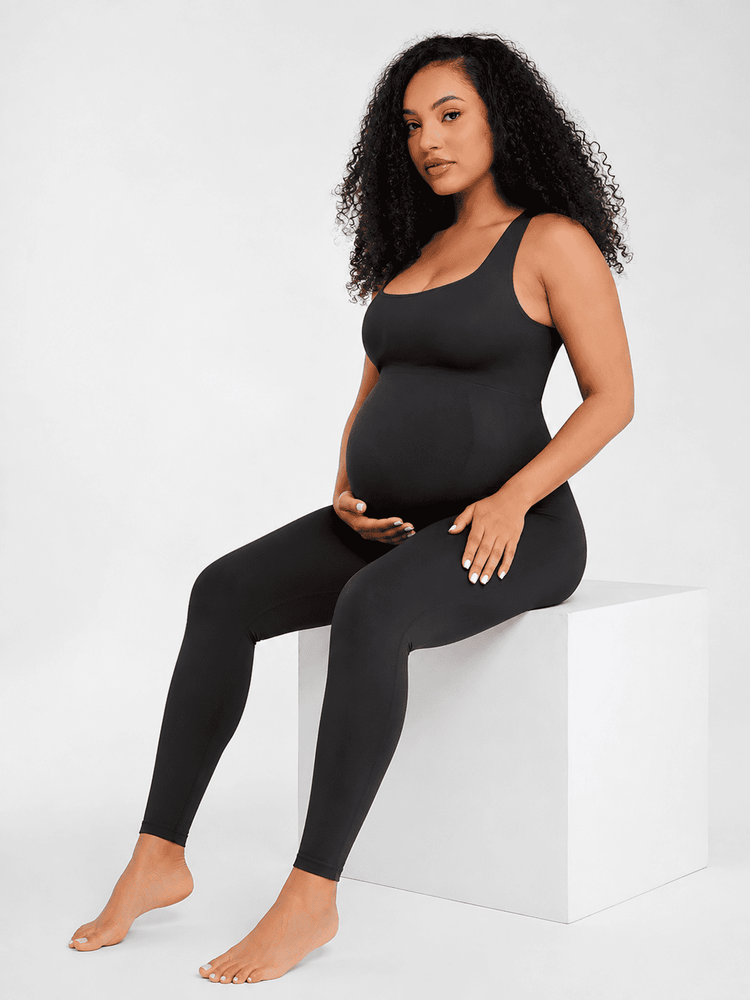 A pregnant model with curly black hair sits on a white block against a light gray studio background, wearing a black maternity tank top and matching black leggings.