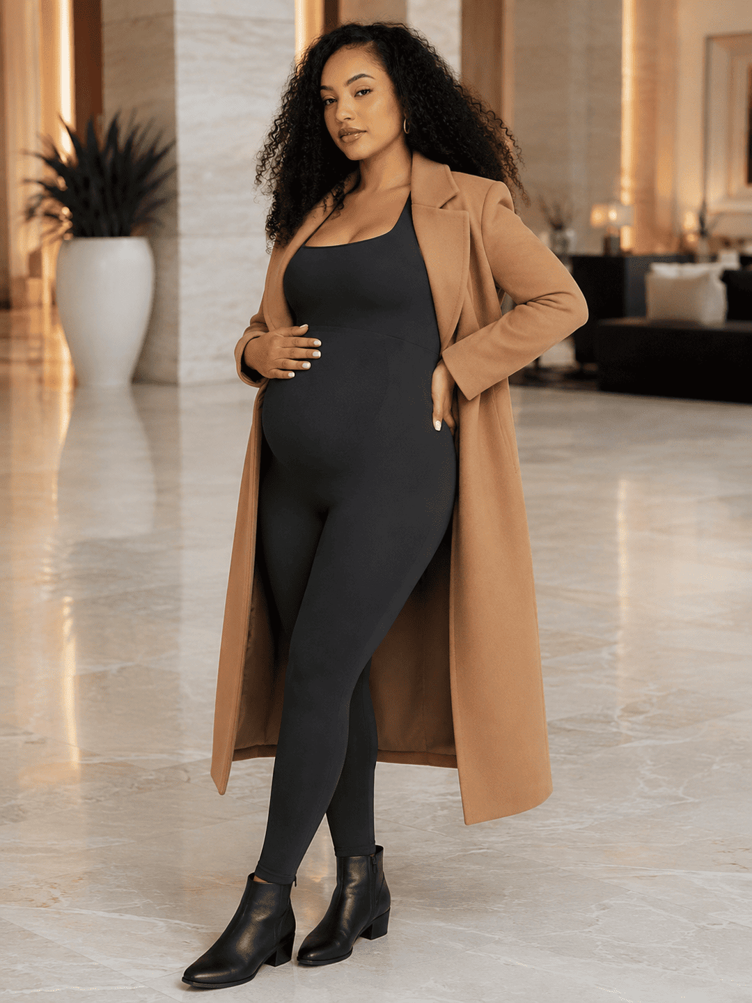 A woman modeling a black maternity bodysuit and long tan coat in a modern, upscale indoor setting with marble floors.