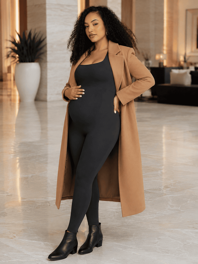 A woman modeling a black maternity bodysuit and long tan coat in a modern, upscale indoor setting with marble floors.