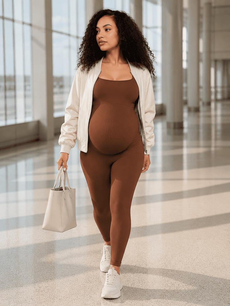 A pregnant woman walking in a bright, modern indoor hallway, wearing a brown bodysuit, a white bomber jacket, and white sneakers, while carrying a light beige tote bag.