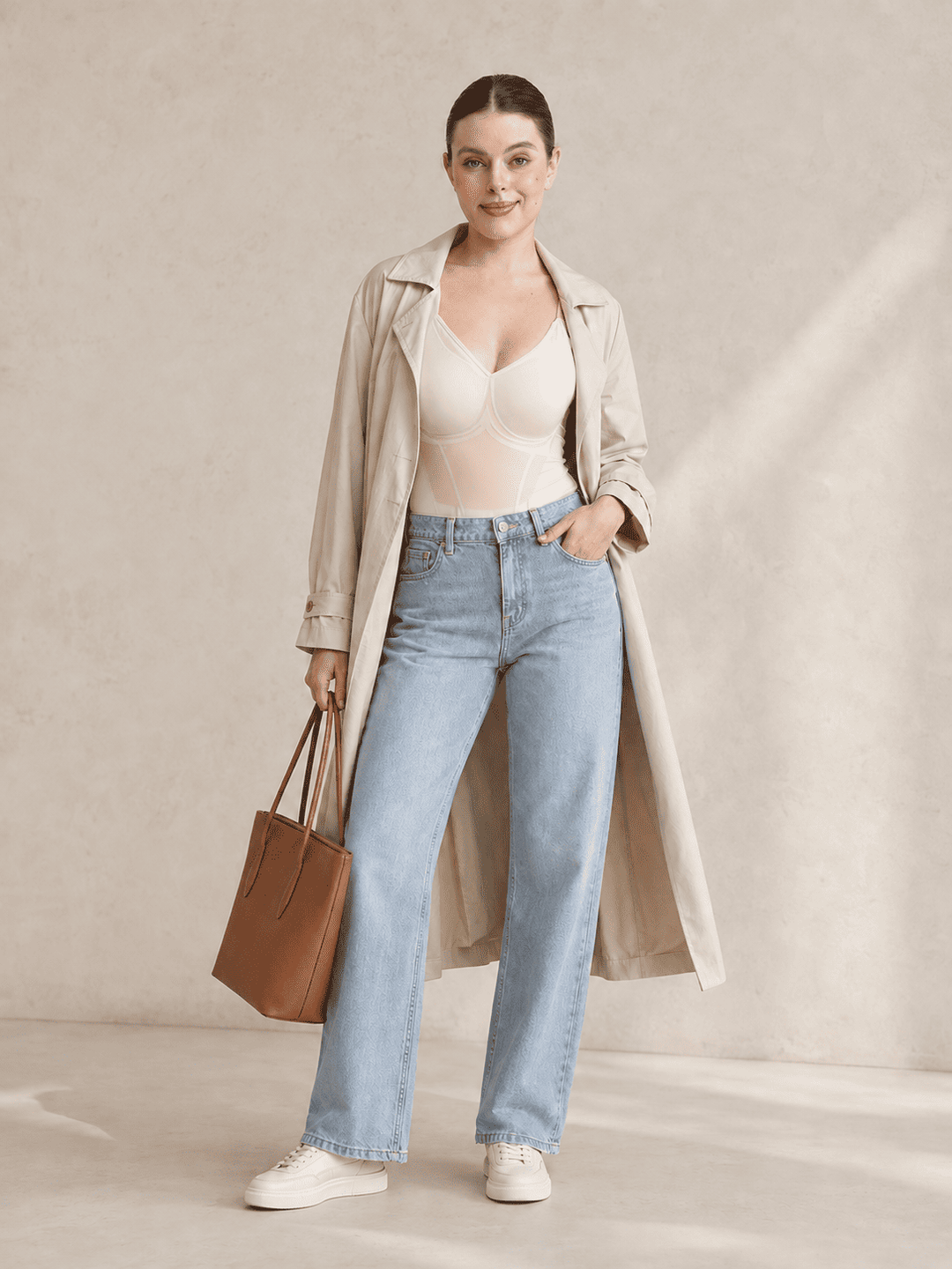 A woman models a light beige trench coat over a beige bodysuit, paired with light-wash blue straight-leg jeans, white sneakers, and a brown leather tote bag, set against a textured beige studio backdrop.