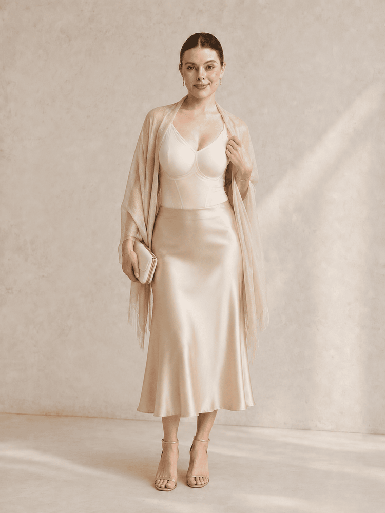 A woman models a champagne-colored satin midi skirt with a matching sheer draped wrap, paired with a structured camisole and nude high-heeled sandals, set against a textured beige studio backdrop.