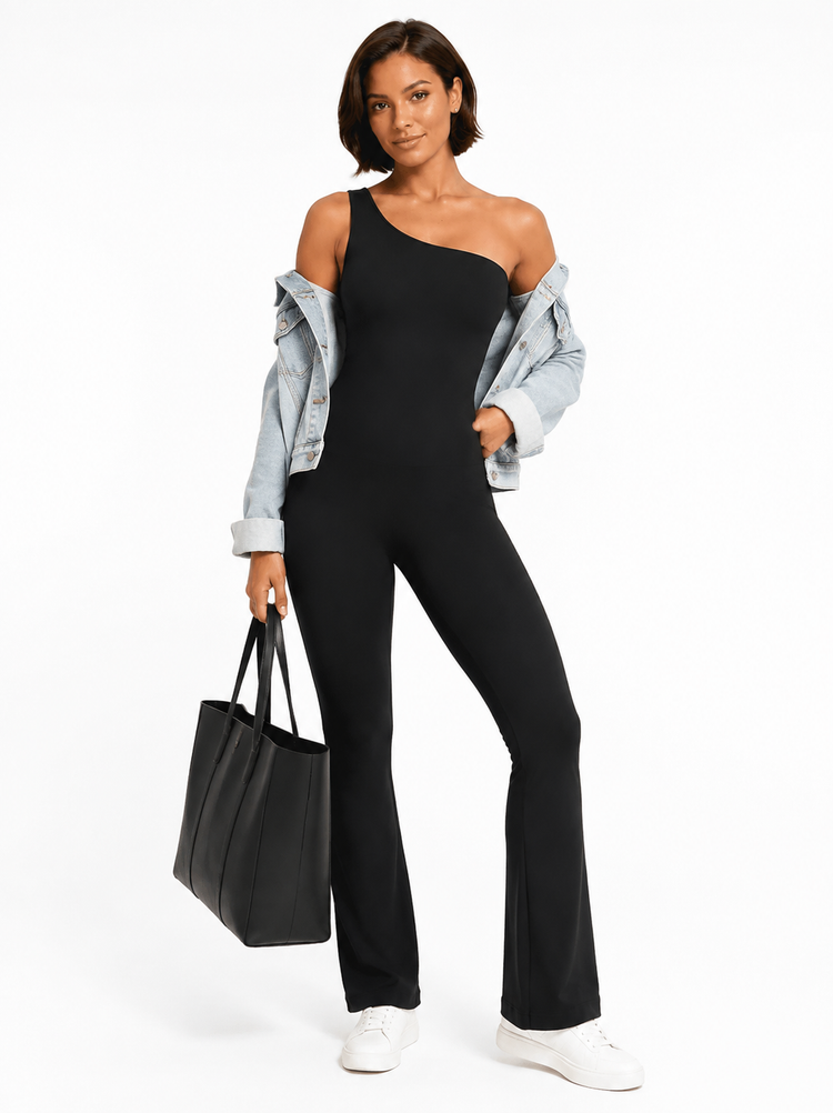 A woman modeling a black one-shoulder flared jumpsuit, styled with an off-the-shoulder light blue denim jacket, white sneakers, and a black tote bag against a plain white studio background.