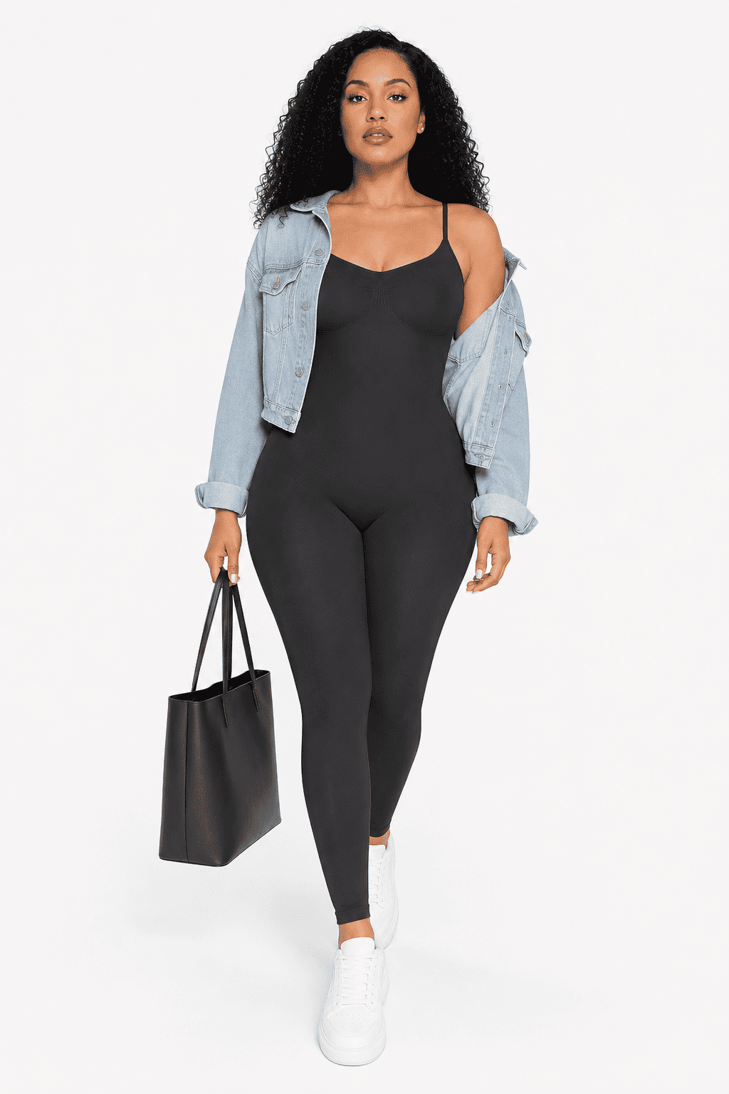 A woman modeling a black camisole jumpsuit with spaghetti straps and full-length legs, styled with an open light-wash denim jacket, white sneakers, and a black tote bag against a plain white studio background.