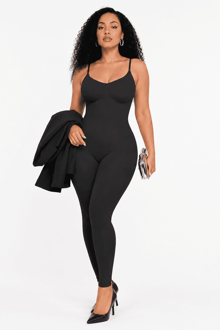 A woman modeling a black spaghetti-strap bodysuit or jumpsuit against a plain white studio background, holding a black blazer under one arm and carrying a small metallic clutch.