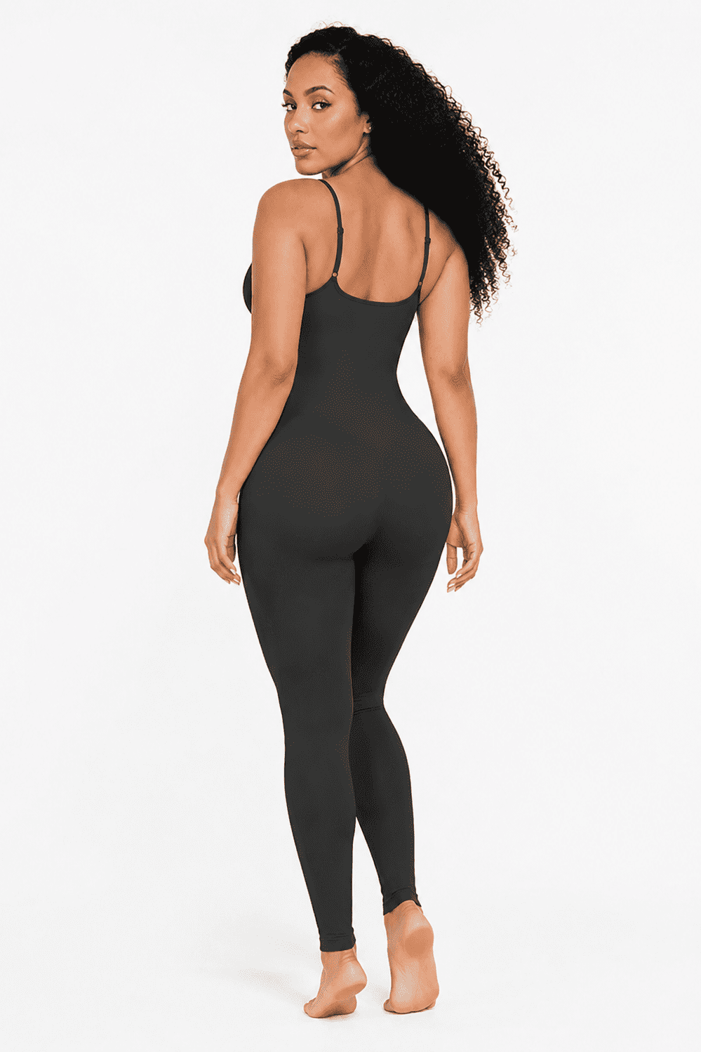 A woman modeling a black full-body shapewear jumpsuit from the back, showing the thin shoulder straps and back design, against a plain white studio background.