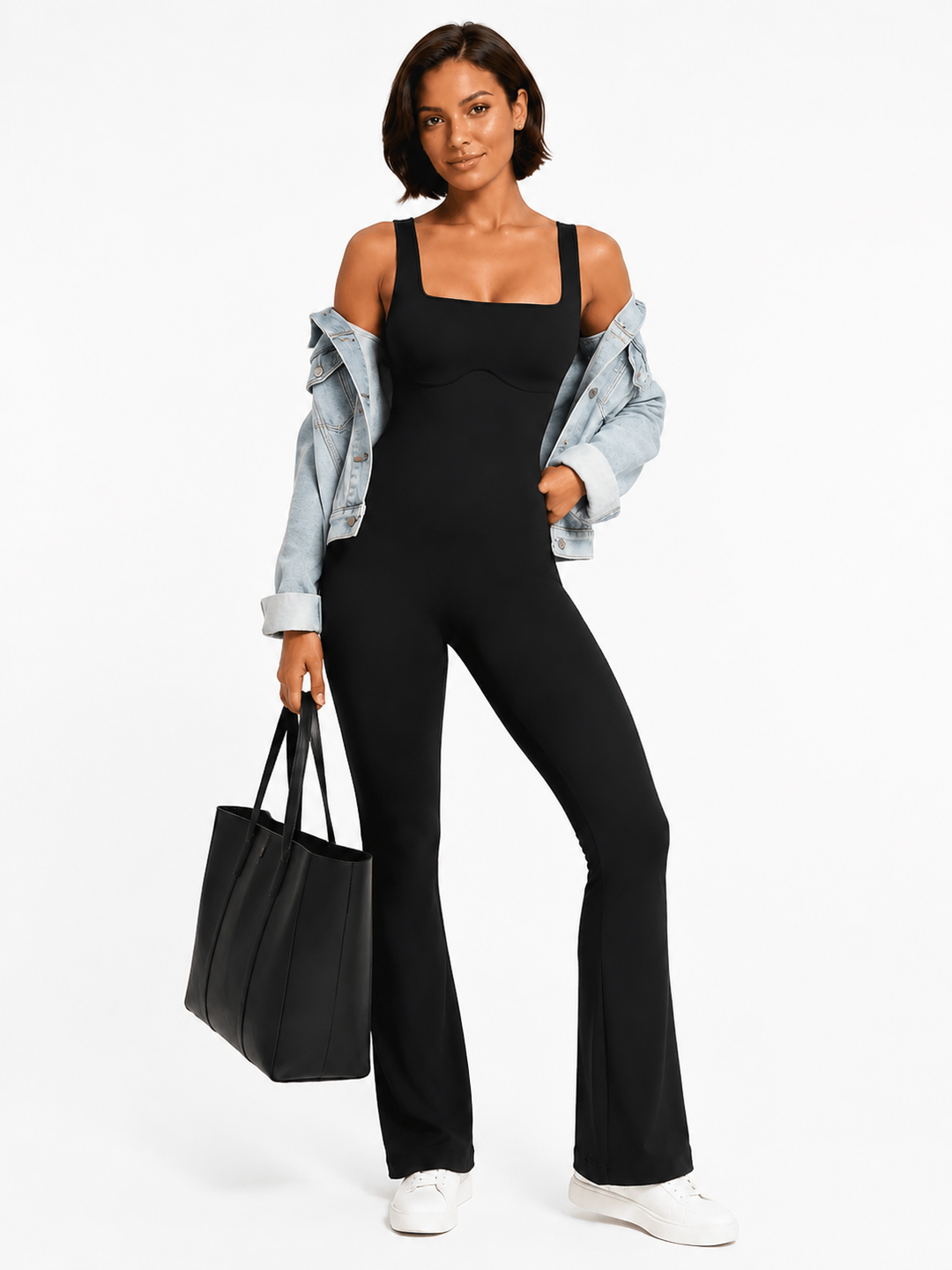 A woman modeling a black sleeveless jumpsuit with a square neckline and wide legs, styled with an open light blue denim jacket, white sneakers, and a black tote bag against a plain white studio background.