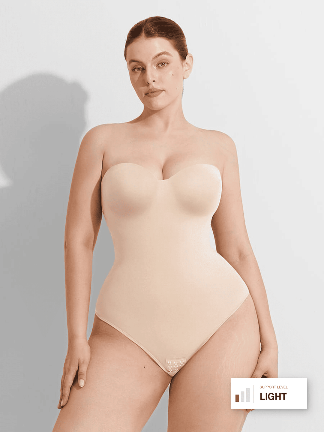 A woman modeling a strapless light beige bodysuit or shapewear garment against a plain light gray studio background. A small label in the lower right reads 'SUPPLEMENTS LEVEL LIGHT'.