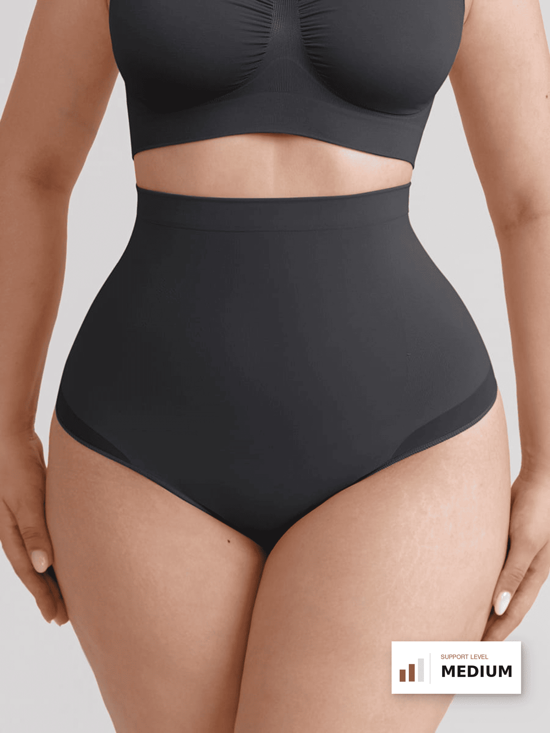 A plus-size model wearing a black high-waisted brief and matching black sports bra against a light gray studio background. A small &