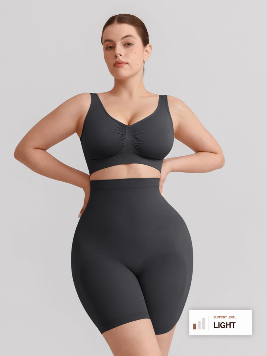 A woman modeling a dark gray two-piece shapewear set consisting of a ruched sports bra and high-waisted shapewear shorts, set against a solid light gray background.