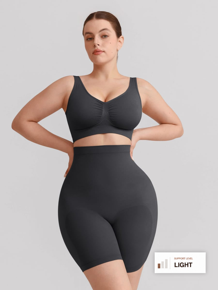 A woman modeling a dark gray two-piece shapewear set consisting of a ruched sports bra and high-waisted shapewear shorts, set against a solid light gray background.