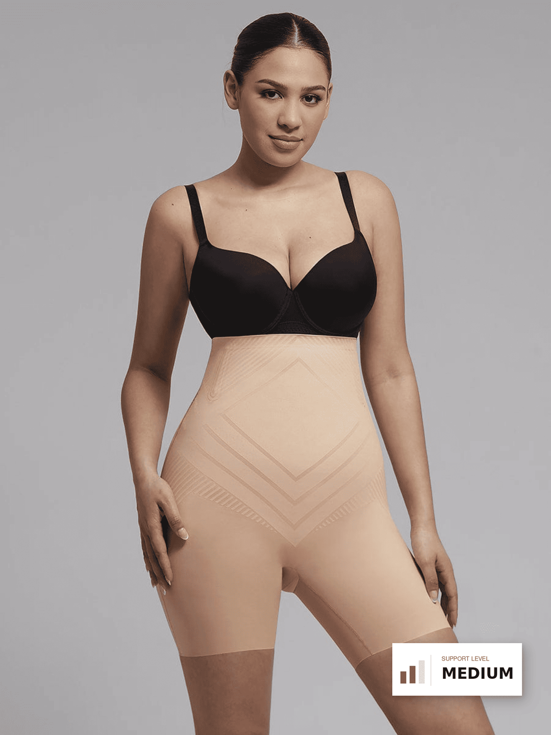 A model wearing a beige high-waisted compression shapewear brief over a black bra, shown against a solid gray studio background with a 'SUPPORT LEVEL MEDIUM' badge in the bottom right corner.