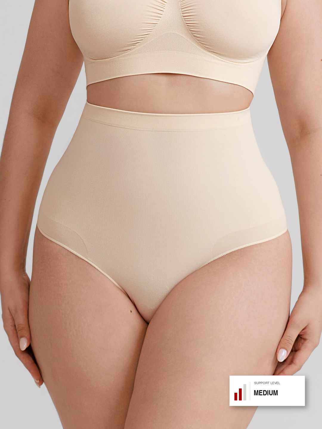 A model wearing a beige high-waisted shapewear brief and matching bralette against a light gray studio background. A small badge in the bottom right corner reads 'SUPPORT LEVEL MEDIUM'.