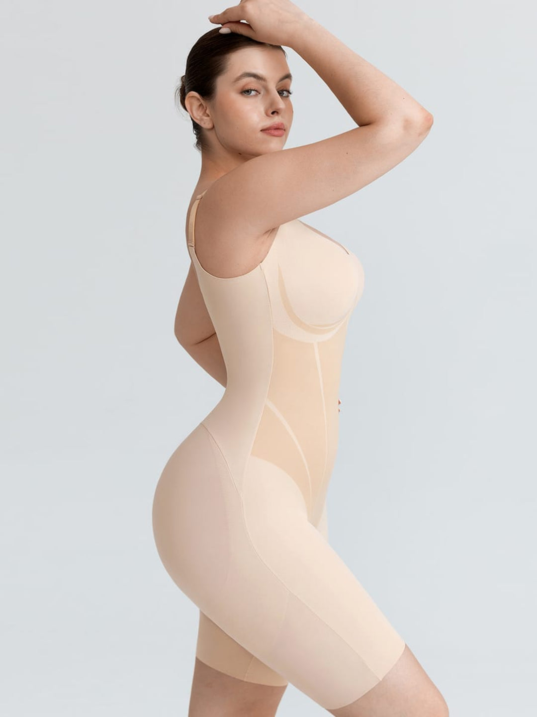 A woman modeling a nude-beige full-body compression shapewear bodysuit against a light gray studio background.