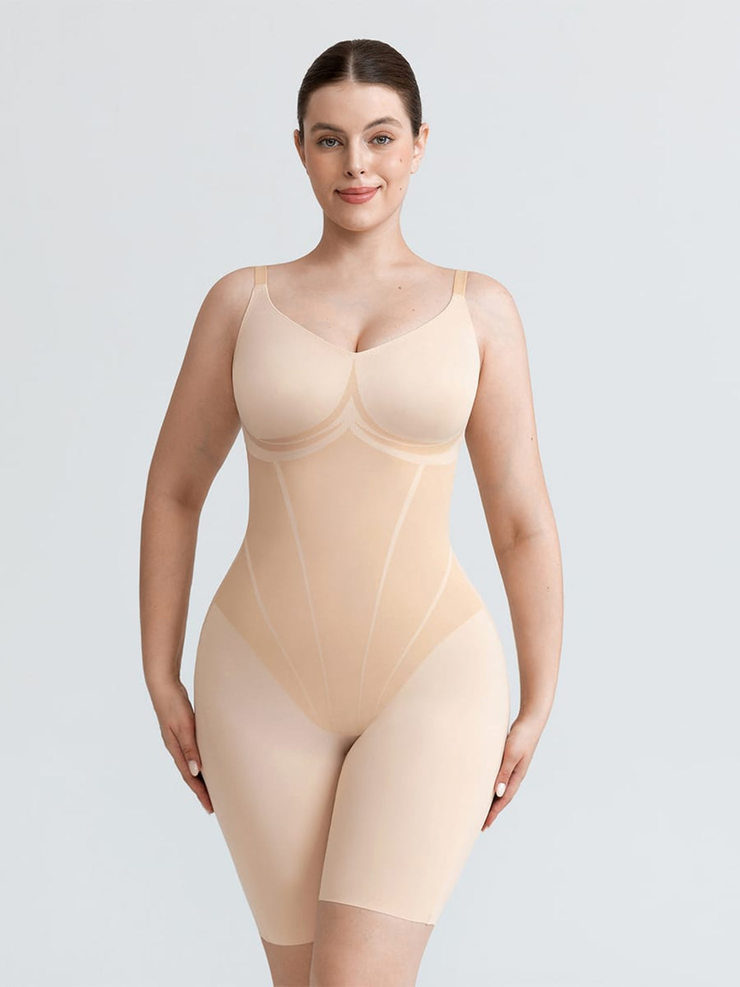 A woman modeling a nude-beige compression bodysuit with built-in bra cups and mid-thigh leggings, shown against a light gray studio background.