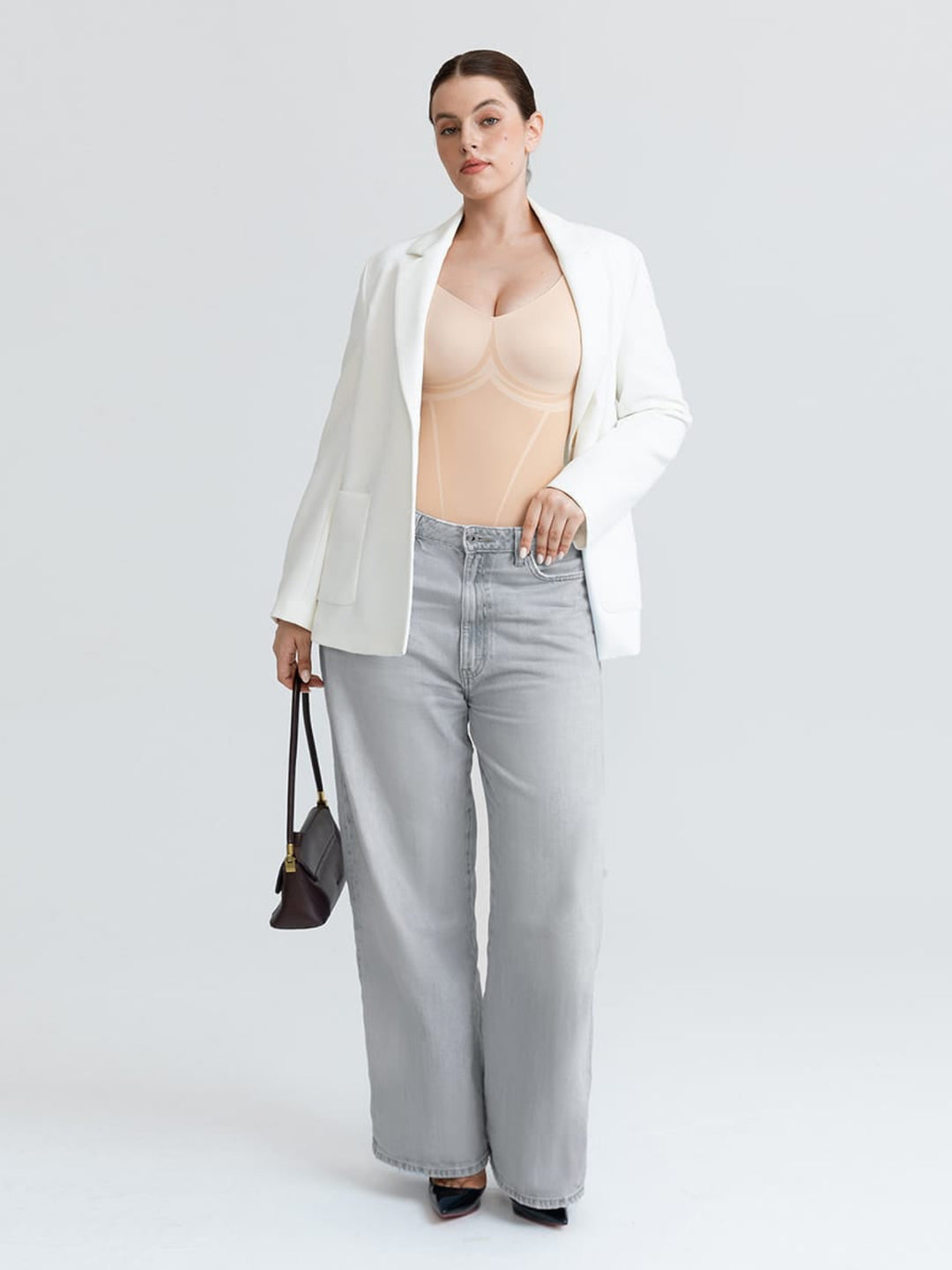 A model stands against a light gray studio background wearing a white blazer, a beige bodysuit, light gray wide-leg jeans, and black heels, while holding a small dark handbag.