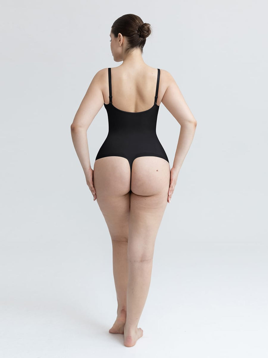 A woman is shown from behind wearing a black bodysuit with thin shoulder straps and a high-cut waist, standing against a plain light gray studio background.