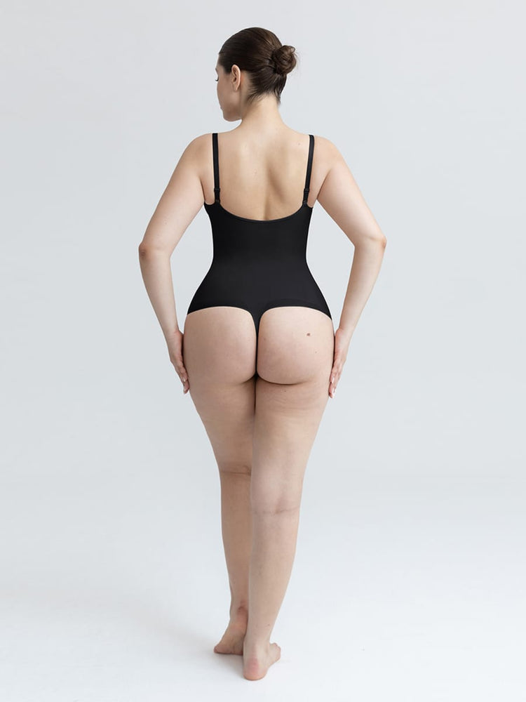 A woman is shown from behind wearing a black bodysuit with thin shoulder straps and a high-cut waist, standing against a plain light gray studio background.