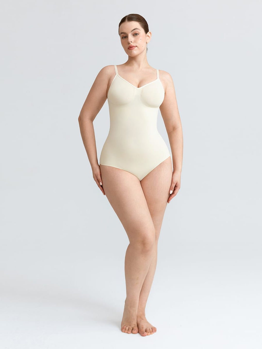 A woman modeling a seamless, skin-toned beige shapewear bodysuit with underwire cups and thin shoulder straps, standing against a light gray studio background.