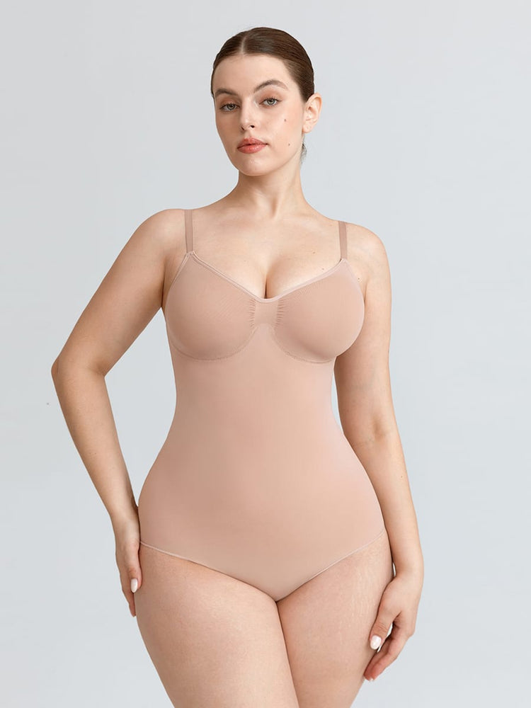 A plus-size model wearing a seamless, light beige bodysuit with underwire cups and matching high-waisted briefs, photographed against a solid light gray studio background.
