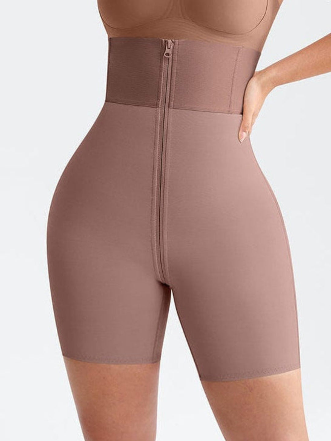 A woman modeling a shapewear bodysuit in a dusty rose-brown color, featuring a bralette top, high-waisted shorts, and a front zipper closure, set against a plain light gray background.