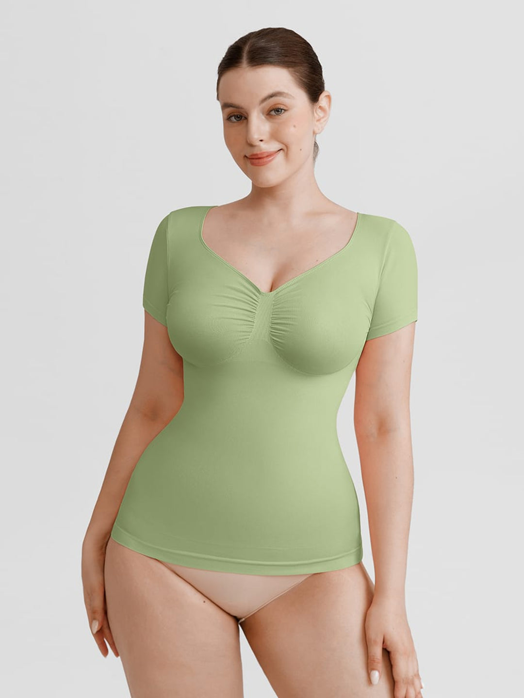 A woman modeling a light green short-sleeve bodysuit with a ruched sweetheart neckline, paired with beige underwear, against a solid light gray background.