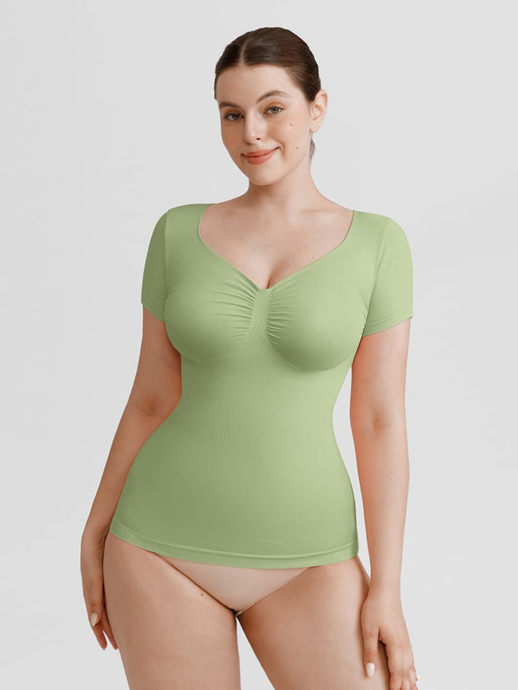 A woman modeling a light green short-sleeve bodysuit with a ruched sweetheart neckline, paired with beige underwear, against a solid light gray background.