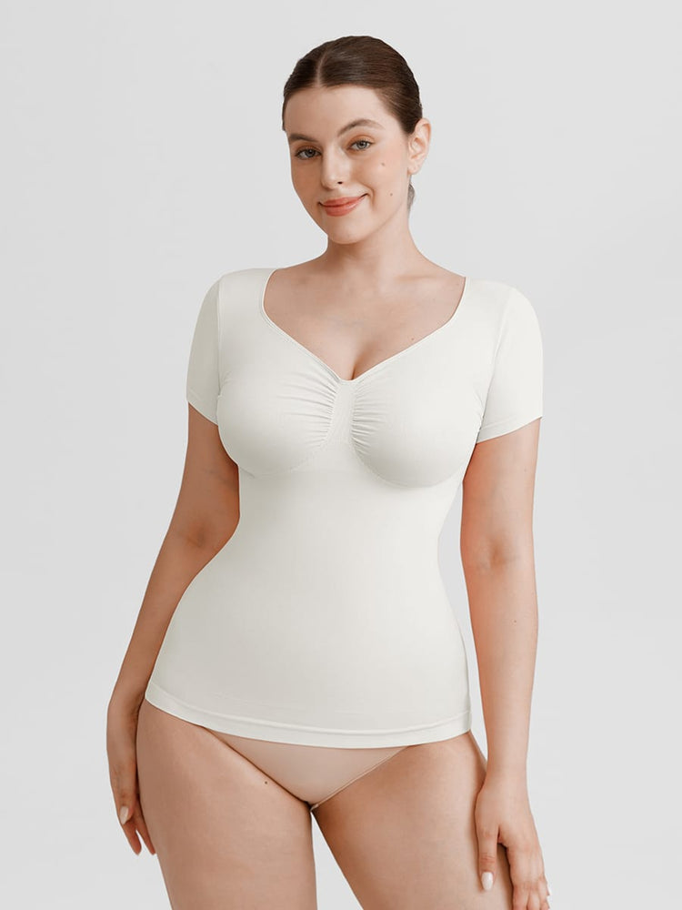 A woman modeling a short-sleeve white bodysuit with a ruched sweetheart neckline, paired with nude high-waisted briefs, against a solid light gray background.