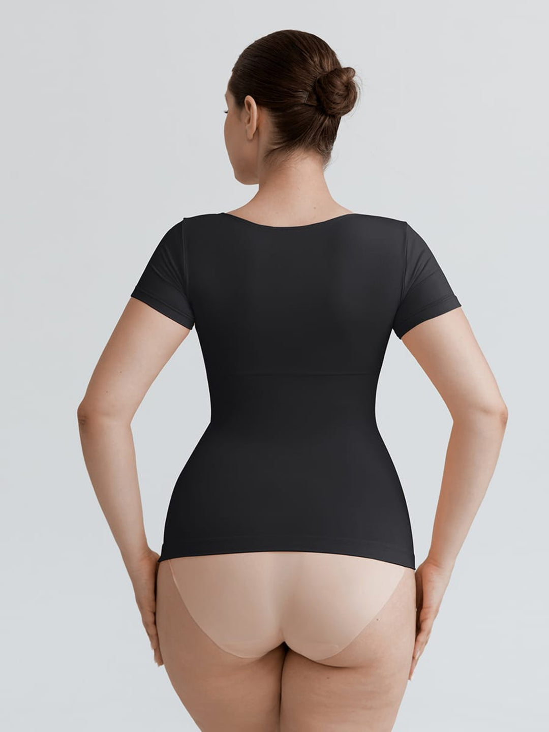 A woman shown from the back wearing a black short-sleeve bodysuit or shapewear top, paired with nude tights, against a light gray studio background.