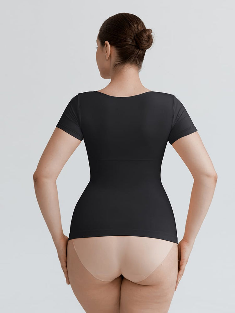 A woman shown from the back wearing a black short-sleeve bodysuit or shapewear top, paired with nude tights, against a light gray studio background.