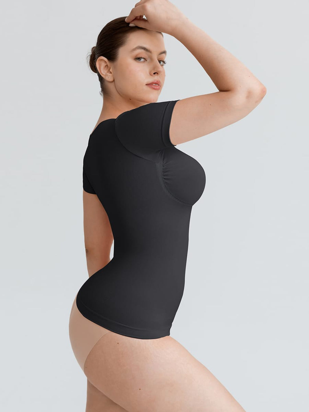 A woman modeling a black short-sleeve bodysuit with structured underbust and bust padding, shown against a plain light gray studio background.